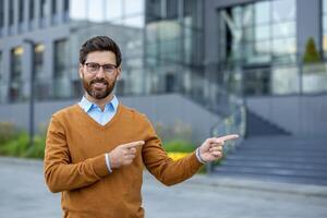 A smiling businessman gestures with both hands, pointing towards the right, near a modern building. He wears glasses and an orange sweater. photo