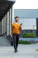 A stylish businessman walks outside, engrossed in his smartphone, carrying a briefcase near a modern building. photo