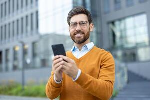 A smiling businessman, holding his phone, stands confidently outside in front of a modern building, showcasing technology. photo