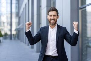A successful businessman celebrates with a fist pump, expressing triumph and joy. He is wearing a suit and stands outside a modern building, looking very happy. photo