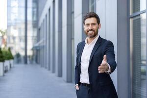 A businessman in a suit offers a handshake, smiling in front of a modern office building. photo