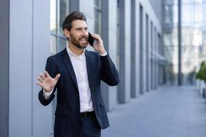 A smiling businessman gestures excitedly while talking on his phone outdoors, near a modern office building. He seems to be in a good mood. photo