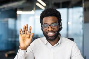 A cheerful man with glasses and a headset waves, expressing a friendly welcome or greeting. He smiles in an office setting. photo
