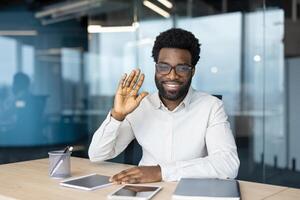 An African-American man in glasses waves and smiles while sitting at a desk in an office. He appears happy and approachable. photo