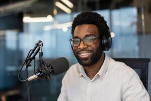 A smiling man with glasses and headphones at a recording studio, speaking into a microphone. photo