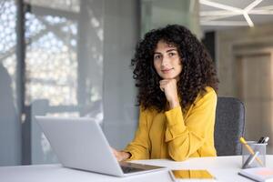 A beautiful woman with curly hair looks at the camera while working on a laptop. She is sitting in an office with a window behind her. photo