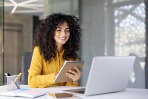 A smiling woman with curly hair takes notes while working on a laptop in a modern office setting. photo