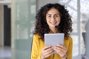 A smiling woman wearing a headset holds a tablet, appearing ready for customer service in a modern office setting. photo