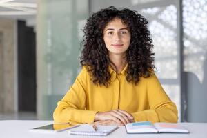 A smiling woman with curly hair sits at a desk with a tablet, notebook and pen in front of her in a bright, modern office space photo