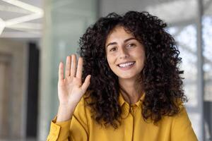 A friendly young woman with curly hair smiles and waves during a conference call, creating a warm and engaging connection photo