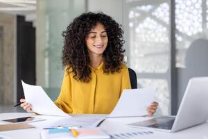 A beautiful businesswoman reviews documents at her office desk, smiling while reading the papers. A laptop, notepad, and tablet are visible. photo