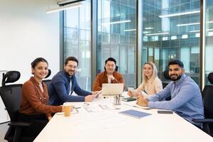 A diverse group of business professionals smiling in a bright, modern office setting, gathered around a meeting table. photo