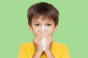 A young boy with a cold, holding a tissue to his nose. He is wearing a yellow shirt and has brown hair. The background is green. T photo