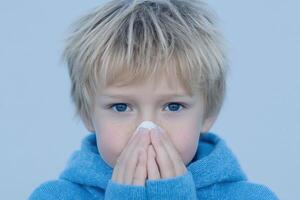A young boy with blond hair blows his nose with a tissue. The image is a portrait of the child's face, showing his blue eyes and photo