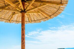 A vibrant, multicolored straw beach umbrella providing shade on a serene sandy beach by the ocean. Clear blue skies and calm waters create a peaceful atmosphere. photo