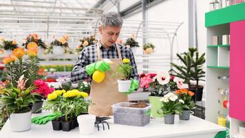 A man wearing gloves and apron, spraying a small plant inside a greenhouse with various flowers. photo