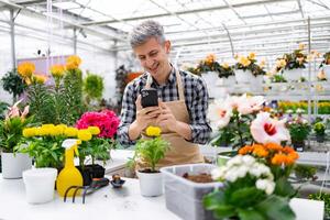 A smiling man in an apron uses his phone while standing in a vibrant greenhouse filled with a variety of colorful flowers and plants. photo