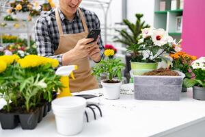 A florist taking a photo of a flower on a phone while surrounded by various plants and gardening tools.