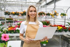 A smiling florist stands in her shop, holding a laptop, with an array of vibrant flowers in the background. photo