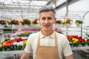 A Caucasian man in a flower shop smiles at the camera, wearing an apron with colorful flowers in the background. photo
