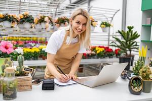 A smiling florist stands at a table, working on a laptop and taking notes in a vibrant shop, surrounded by various flowers and plants. photo