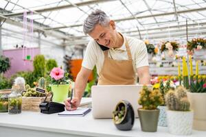 A Caucasian man in a florist shop works, talking on the phone, writing in a notebook, and using a laptop at the counter. photo