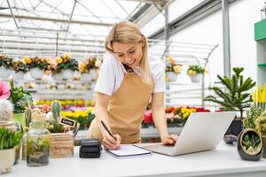 A Caucasian woman takes an order at a florist shop, using a laptop and a phone. She is writing down the details while surrounded by various plants. photo