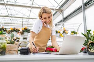 A Caucasian woman working in a florist shop, taking a call and using a laptop while writing. photo