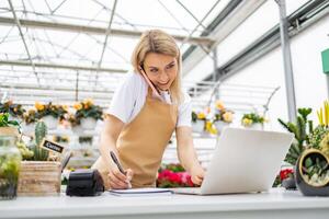 A Caucasian woman, working at a flower shop, is on the phone, taking notes, and using a laptop on a bright day. photo