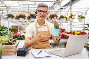 A Caucasian man in a florist shop holds a cactus in a pot while on a call, smiling at the camera and gesturing. photo