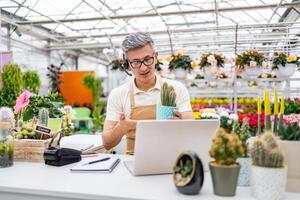 A Caucasian florist assisting a customer online in a greenhouse setting, showcasing a cactus on a call. photo