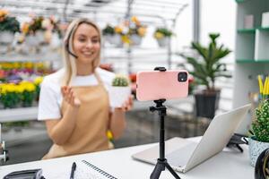 A Caucasian woman in a florist shop hosts a live sale of a cactus, using her smartphone for a broadcast. photo