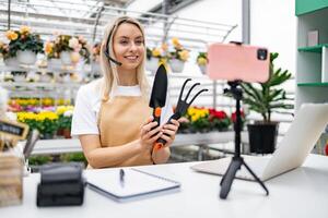A Caucasian woman, working in a florist shop, presents gardening tools while streaming online from a table with a laptop and smartphone. photo