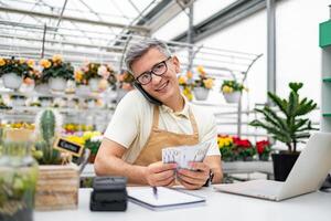 A Caucasian florist, happy with money in hand, smiles while on a phone call in his shop, surrounded by plants and flowers. photo