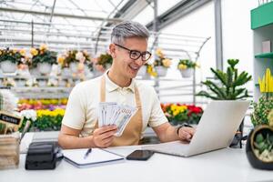 A Caucasian man, a florist, enjoys success at a sales table in his shop, counting money after a profitable day with a laptop. photo