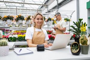 A florist is at work, smiling and using a laptop. In the background, a colleague is inspecting a plant, in a sunny greenhouse filled with flowers. photo