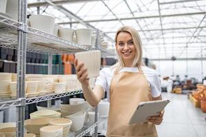 A blonde Caucasian woman in an apron selects a flowerpot in a shop. She is holding a tablet, smiling at the viewer, showcasing various plant containers. photo