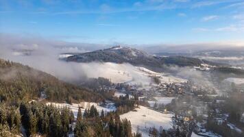 Aerial view captures the snowy terrain surrounding Tanvaldsky Spicak in Czechia. Fog drifts over hills while the village nestles beneath the snow-capped peaks under a clear sky. photo