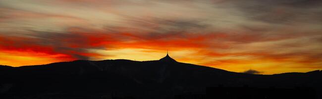 The sky over Jested Mountain transforms into a canvas of warm colors during winter evenings. The silhouette of the mountain contrasts beautifully against the vibrant sunset sky. photo