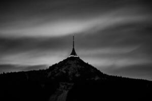 Jested Mountain stands majestically against a winter evening sky in Czechia. The tower at its peak is illuminated, contrasting with the darkened landscape and clouds. Black and white photography photo