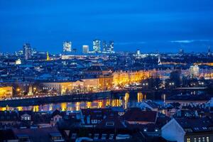The National Theatre stands illuminated against the evening sky in Prague, with modern buildings of Pankrac rising in the background. Reflections shimmer in the water. photo