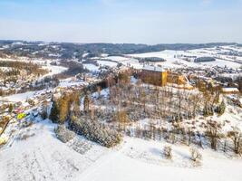 Pecka medieval castle stands proudly atop a hill, blanketed in snow. The surrounding landscape features frosted trees and quiet village below, creating a picturesque winter setting. photo