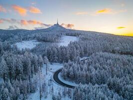 A stunning winter twilight view of Jested in Czechia, showcasing snow-covered trees and a winding road leading up to the iconic tower. The colors of sunset enhance the serene atmosphere. photo