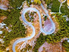 A serpentine road meanders through a vibrant forest landscape during autumn. The surrounding trees showcase rich colors, creating a picturesque view from above. photo