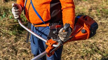 A person using a string trimmer to cut grass in a yard. Wearing gloves and an orange shirt, they focus on lawn maintenance, representing landscaping and gardening tasks. photo