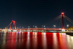 A stunning night view of the illuminated Willemsbrug Bridge in Rotterdam, Netherlands, with beautiful long exposure light trails creating a mesmerizing effect. photo