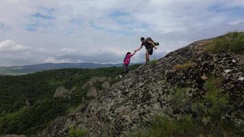 aerial view close upMan helps woman climb rocky trail on mountain slope with lush forest and cloudy sky in background, teamwork and adventure concept video