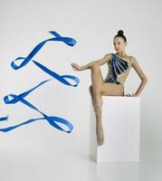 a girl from rhythmic gymnastics in a bodysuit rotates a ribbon while sitting on a cube photo