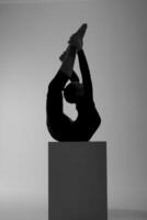 the silhouette of a girl from rhythmic gymnastics in a bodysuit on a cube in a deflection holds her feet with her hands photo