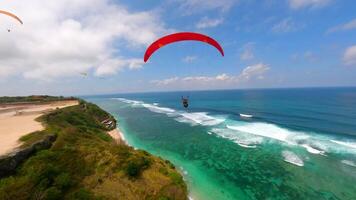 Cinematic FPV drone shot of paragliders gliding over cliffs and turquoise ocean at Pandawa Beach, Bali. Aerial view of dramatic coastline, tropical waves, and summer sky. video